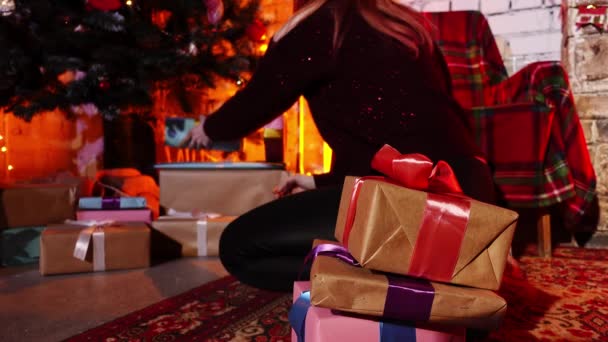 Blanke dame zit op de vloer naast een versierde kerstboom en verbergt er geschenkdozen onder. — Stockvideo