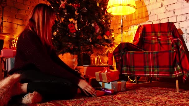 装飾されたクリスマスツリーの隣の床に座っている若い女性とそれの下にギフトボックスを隠す — ストック動画