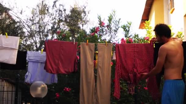 A topless man in blue shorts is taking off clean clothes from the rope and putting them into basket — 图库视频影像