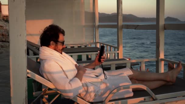 Jonge man met baard praat via video op zijn smartphone liggend op een zonnebank in de lounge — Stockvideo