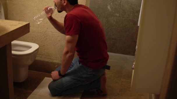 En sjuk man sitter framför toaletten och dricker rent vatten ur flaskan efter att ha kräkts — Stockvideo