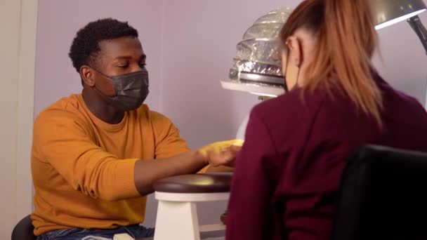 Αφρικανός με προστατευτική μάσκα σε ινστιτούτο ομορφιάς φροντίζει τα νύχια ενός μανικιούρ. — Αρχείο Βίντεο