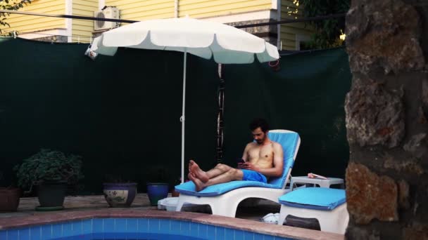 Young man is resting next to the swimming pool on a lounge chair and using smartphone in his hands — Stockvideo