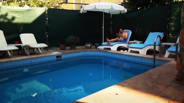 A male tourist is having rest on lounge chair near the swimming pool chatting to someone — Vídeo de Stock