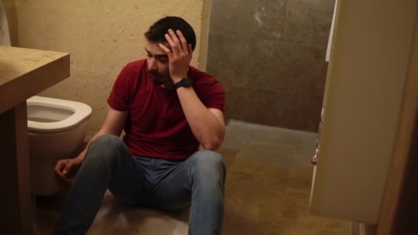 Genç sakallı sarhoş adam tuvaletin yanında oturuyor karnını tutuyor ve saf su içiyor. — Stok video