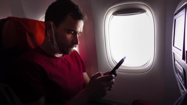 वैद्यकीय संरक्षक मास्कमध्ये एक तरुण माणूस विमानात फोनवर बोलत आहे — स्टॉक व्हिडिओ