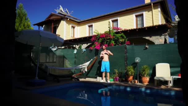Young caucasian man is applying sunscreen lotion on his slim body standing next to the swimming pool — Vídeo de Stock
