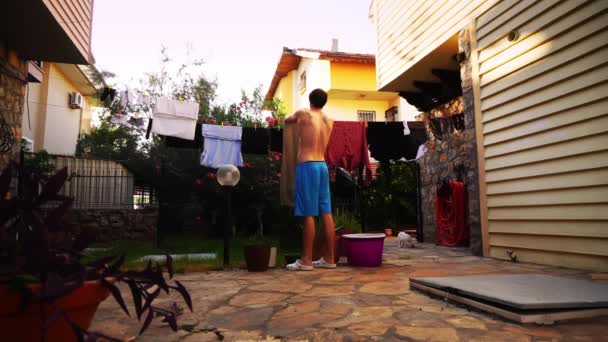 A man in swimwear is hanging wet clean clothes on clothesline on the backyard of guest house — Vídeo de Stock