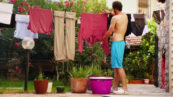 Homme en short vient à l'arrière-cour de la villa pour enlever des vêtements propres et humides d'une ligne — Video