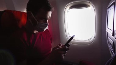 Tıbbi maskeli bir adam uçağın penceresinin yanında oturuyor ve akıllı telefonuna mesaj atıyor.