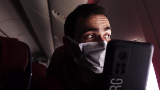 El hombre con máscara médica está hablando por videollamada usando un teléfono inteligente y sentado en el avión — Vídeos de Stock
