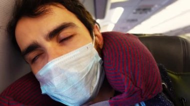Tıbbi koruyucu maskeli yorgun genç adam uçakta uyumaya çalışıyor.