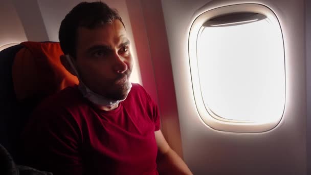 비행기 안에 앉아 있는 남성 승객은 의료용 마스크를 쓴 채 비행기 창문 옆에 앉아 턱을 잡아당긴다 — 비디오