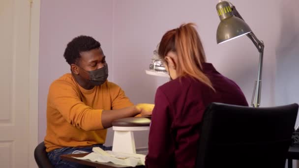 Mujer joven está haciendo manicura al hombre negro con máscara en el salón de spa siguiendo las reglas de seguridad — Vídeos de Stock