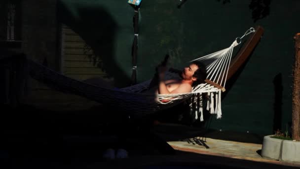 Giovane uomo caucasico sta prendendo il sole sdraiato sull'amaca e utilizzando smartphone nelle sue mani — Video Stock