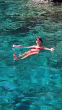 Genç bir kadın köpüklü erişteyle denizde yüzüyor. Gözleri ağır çekimde kapalı.