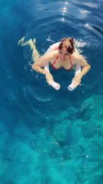 Mavi, berrak bir denizde köpüklü erişteyle yüzen neşeli genç bir kadının dikey çekimi.