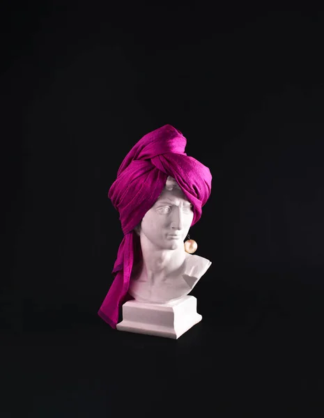 Plaster male head wrapped with hot pink turban against dark background. Popular culture concept. Man with pearl earing creative layout. Mockery, consumerism, irony. Bad hair day ridiculous idea.