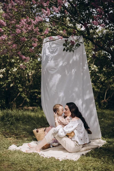 Piknik battaniyesinde oturup bebeğine sarılan mutlu genç bir kadın. Anne ve kızının yaz pikniği leylak bahçesinde. Genç esmer anne ve kızı..