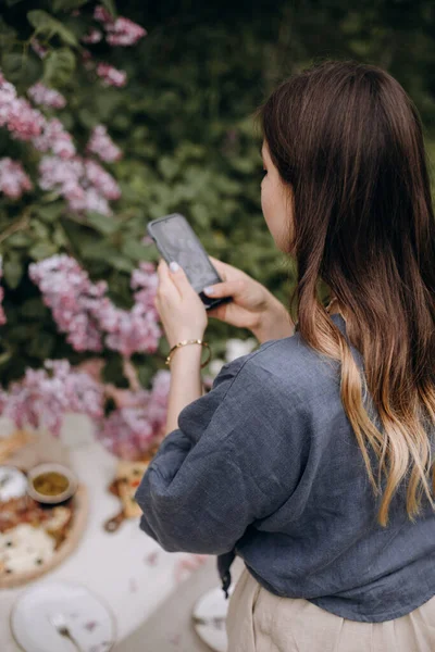 少女は電話でピクニック写真を撮る 波状の髪をした若いブルネットの女性が公園の電話で写真を撮る ストックフォト