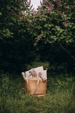bag with a set for a summer picnic. straw bag with wine bottle, glasses and ass pillows. bag with picnic items on the lawn in the lilac garden.