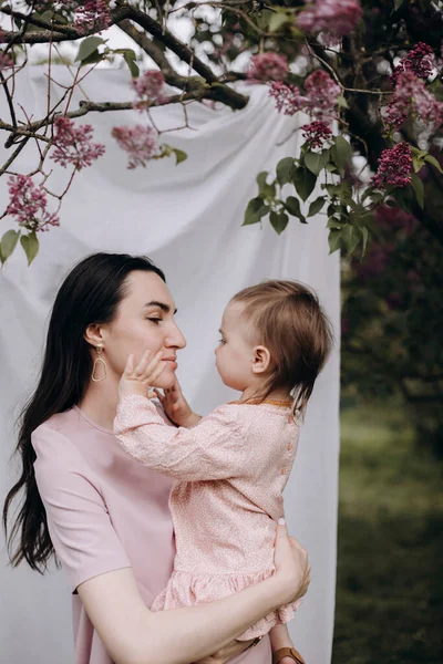 黑发妈妈把她的女婴抱在外面 年轻的母亲抱着她的孩子 这孩子是用母亲的脸玩耍的 在白色光唱机旁边的蓝色花园里带着孩子的母亲 — 图库照片