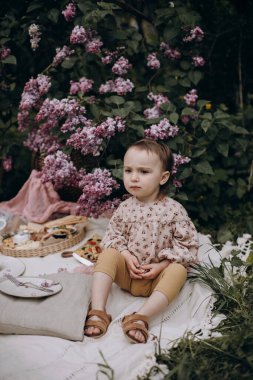 Ciddi bakışlı küçük kız parkta ya da bahçede piknik battaniyesine oturuyor. Piknikte leylak çalılarının yanında oturan bir kız. Çocuk bir şey düşünüyor. Çocuk ailesini bekliyor.