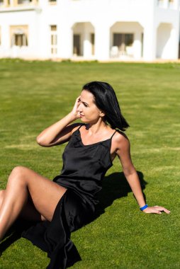Siyah elbiseli genç ve güzel bir kadın yeşil çimlerde oturuyor.