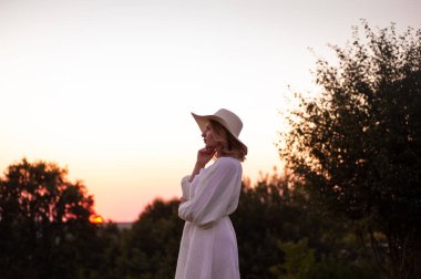 beautiful young woman in a hat and a white dress on a background of sunset