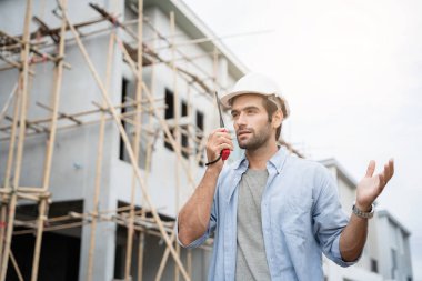 İnşaat alanında duran genç erkek mühendisler taşınabilir telsizle konuşuyorlar ve çeşitli fonksiyonları kontrol ediyorlar..