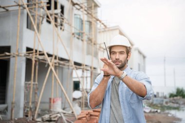 İnşaat alanında duran genç erkek mühendisler taşınabilir telsizle konuşuyorlar ve çeşitli fonksiyonları kontrol ediyorlar..