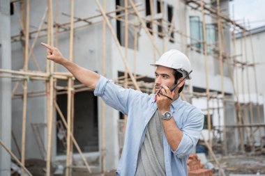 İnşaat alanında duran genç erkek mühendisler taşınabilir telsizle konuşuyorlar ve çeşitli fonksiyonları kontrol ediyorlar..