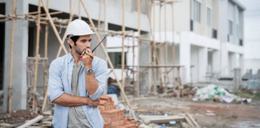 İnşaat alanında duran genç erkek mühendisler taşınabilir telsizle konuşuyorlar ve çeşitli fonksiyonları kontrol ediyorlar..