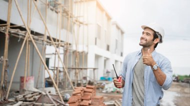İnşaat alanında duran genç erkek mühendisler taşınabilir telsizle konuşuyorlar ve çeşitli fonksiyonları kontrol ediyorlar..