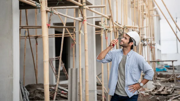 İnşaat alanında duran genç erkek mühendisler taşınabilir telsizle konuşuyorlar ve çeşitli fonksiyonları kontrol ediyorlar..