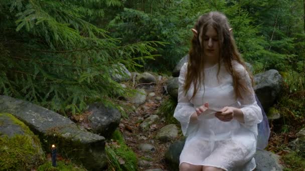 Эльфийская девушка в белом платье открывает конверт с письмами в лесу — стоковое видео