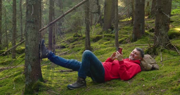 Туристичний чоловік з телефоном відпочиває на моху в прекрасному лісі — стокове відео