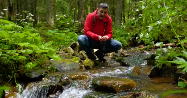 Бородатий чоловік з рюкзаком миється водою з гірської течії — стокове відео