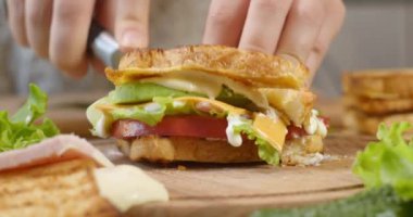 Kadın aşçı, kulüp sandviçini, mutfak tahtasındaki keskin bir bıçakla ikiye bölüyor.