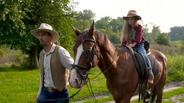 A young cowboy is leading a horse on which his daughter is sitting — Stock Video