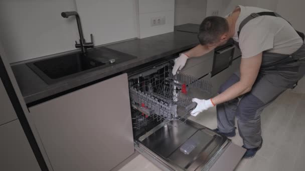 Um reparador profissional em macacão verifica uma máquina de lavar louça doméstica na cozinha — Vídeo de Stock