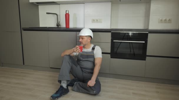 O trabalhador se senta no chão, descansando e bebendo chá na nova cozinha. — Vídeo de Stock