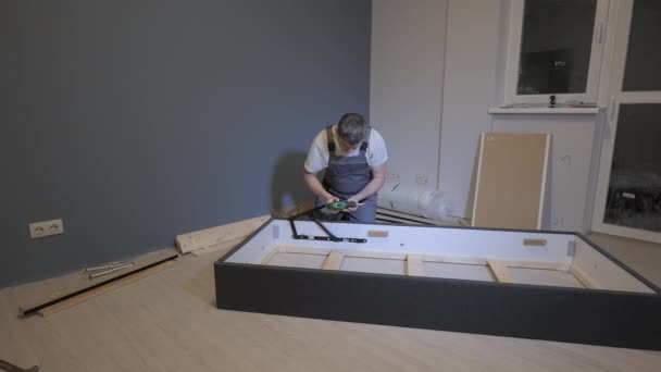 Un constructor en un mono de trabajo está montando una cama en un nuevo apartamento — Vídeos de Stock