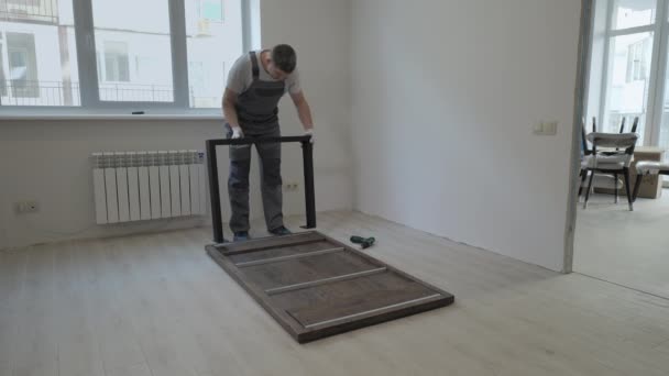 Строитель в рабочем комбинезоне собирает стол в новой квартире — стоковое видео