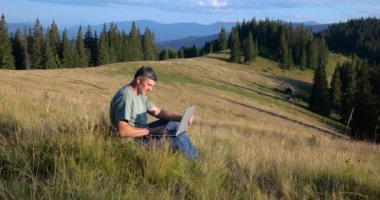 A man sits on a beautiful meadow in the mountains, works on a laptop. Concept of freelancing, digital nomad or remote office.