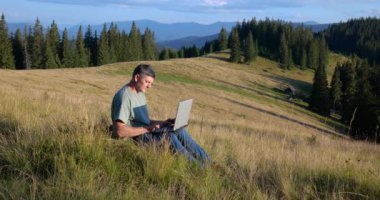 A man sits on a beautiful meadow in the mountains, works on a laptop. Concept of freelancing, digital nomad or remote office.