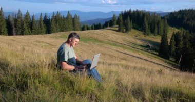 A man sits on a beautiful meadow in the mountains, works on a laptop. Concept of freelancing, digital nomad or remote office.
