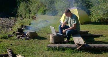 A man works on a laptop in a tourist camp in a beautiful forest. Concept of freelancing, digital nomad or remote office. 4K
