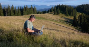 A man sits on a beautiful meadow in the mountains, works on a laptop. Concept of freelancing, digital nomad or remote office.