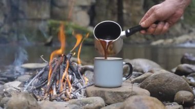 Coffee is poured into a mug from a coffee maker. Breakfast near the fire on the bank of a beautiful river. Travel concept. 4k